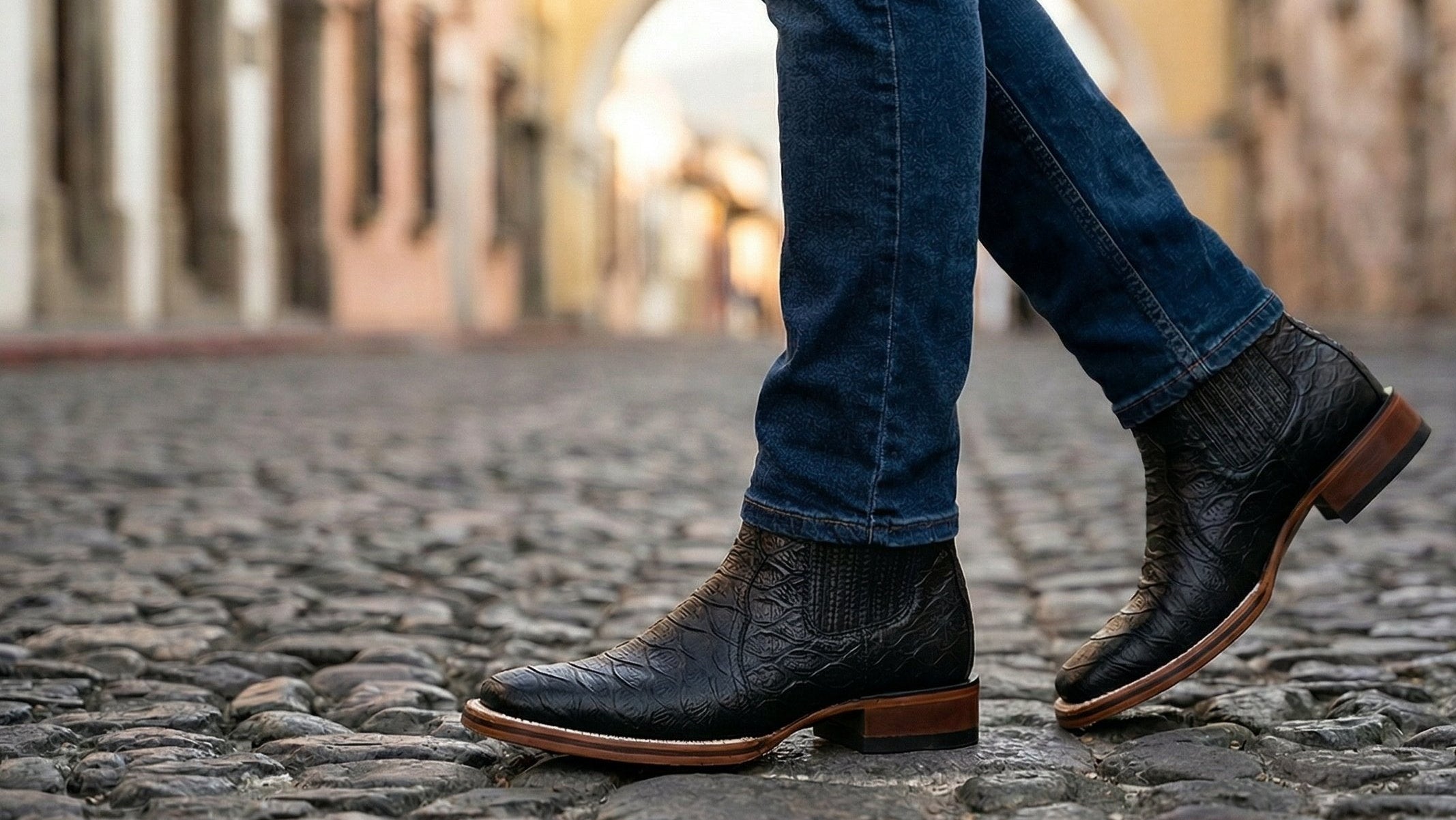 Person wearing black leather shoes and blue jeans walking on a cobblestone street.