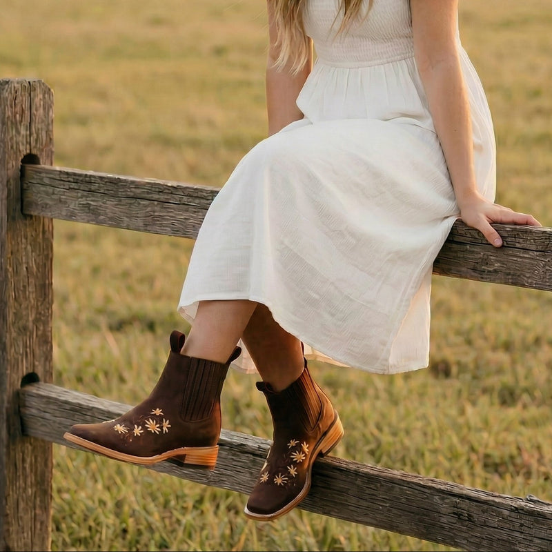 Woman in a white dress and brown hat sitting on a wooden fence in a field.